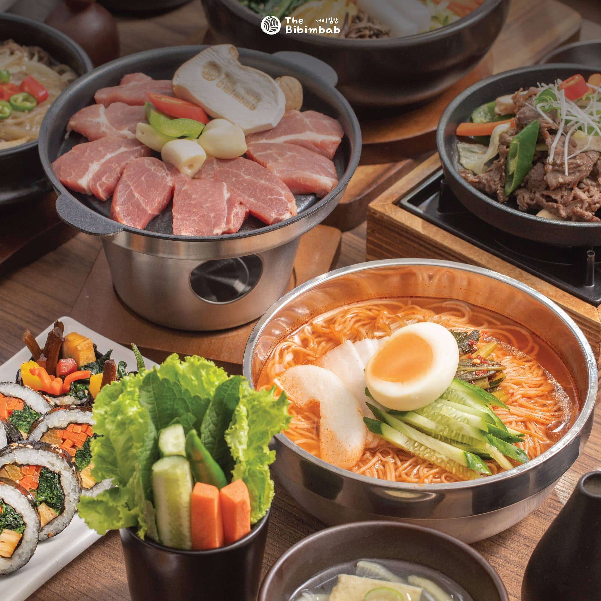 A Korean table with a griddle pan of pork, cold noodles, kimbap and wrapping vegetables
